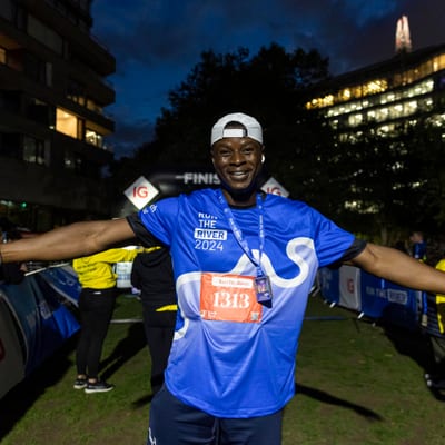 A smiling runner wearing a blue "Run the River 2024" shirt and race bib 1313 stands with arms outstretched at a nighttime event near the finish line, surrounded by people and city buildings.