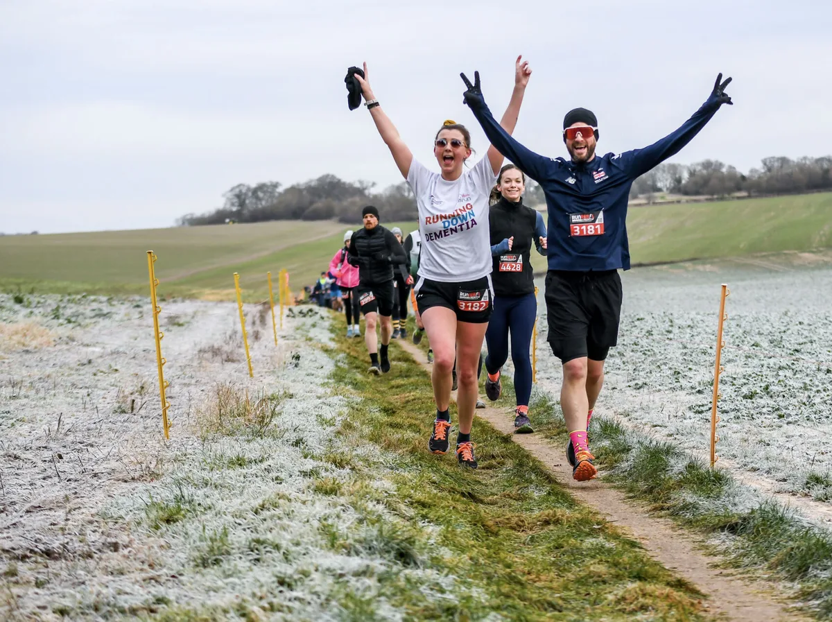 A group of runners on a frosty, grassy trail. Two people in front, smiling with arms raised in celebration, lead others down the path. The surrounding fields appear lightly dusted with frost.