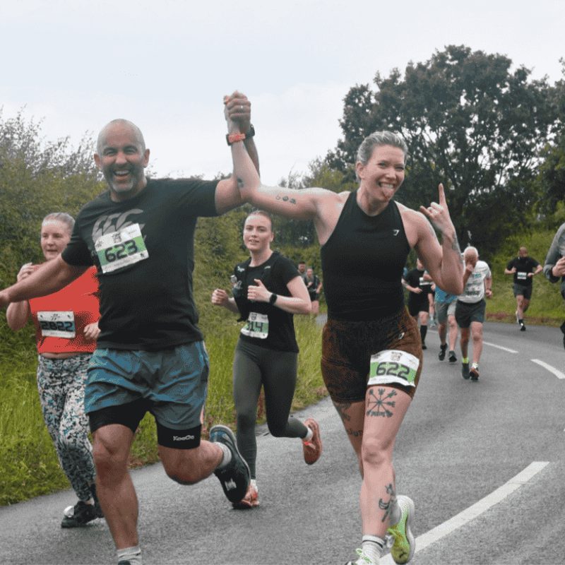 Runners participate in a race on a rural road. A man and woman hold hands, smiling and celebrating as they lead the group. Lush greenery and bushes line the road on both sides. Other runners follow behind, some focused and others cheerful.