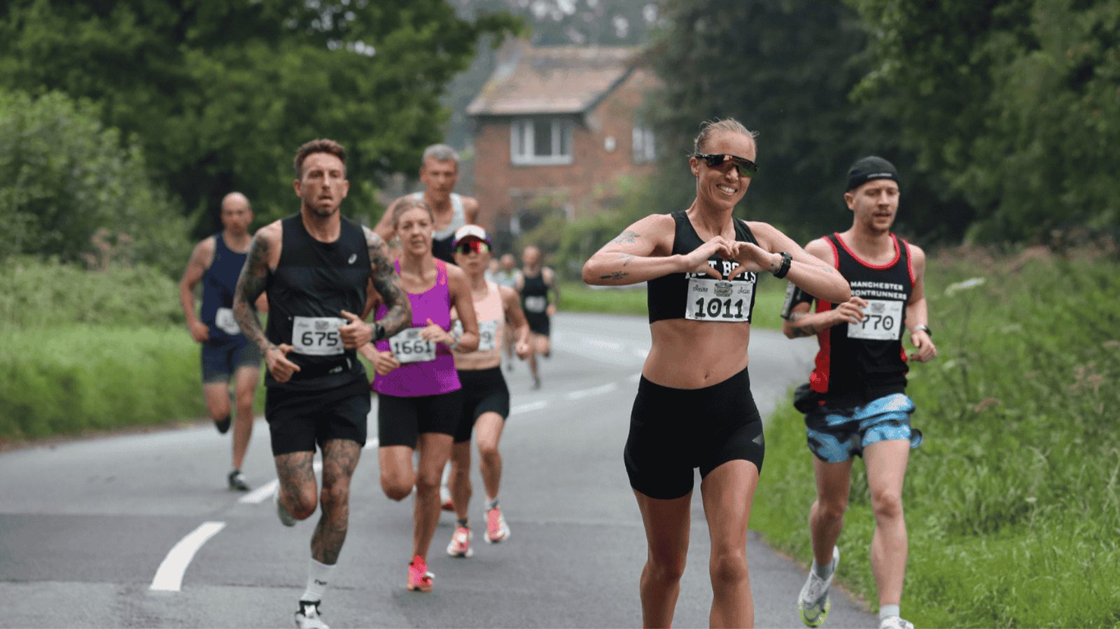 A group of runners participating in a race on a rural road, flanked by greenery. One woman in the foreground is smiling and gesturing with her hands. Participants wear numbered bibs and a variety of athletic gear.