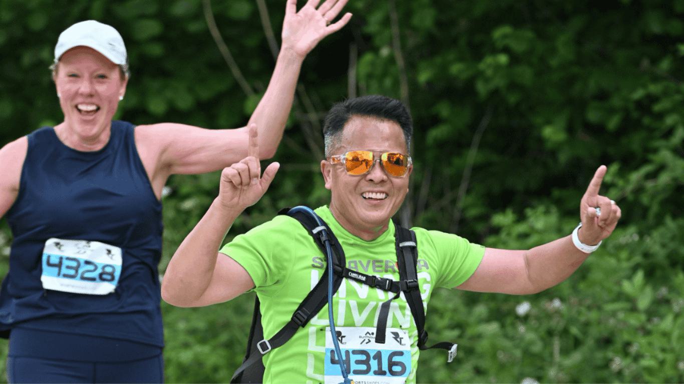 A man and a woman are running outdoors, smiling and waving. The man is wearing orange reflective sunglasses and a green shirt with the number 4316. The woman is wearing a navy tank top with the number 4328. Green foliage is in the background.