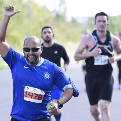 A man in a blue sports shirt runs and raises his hand in a thumbs-up gesture during a race. Other runners are visible in the background on a paved path lined with greenery.
