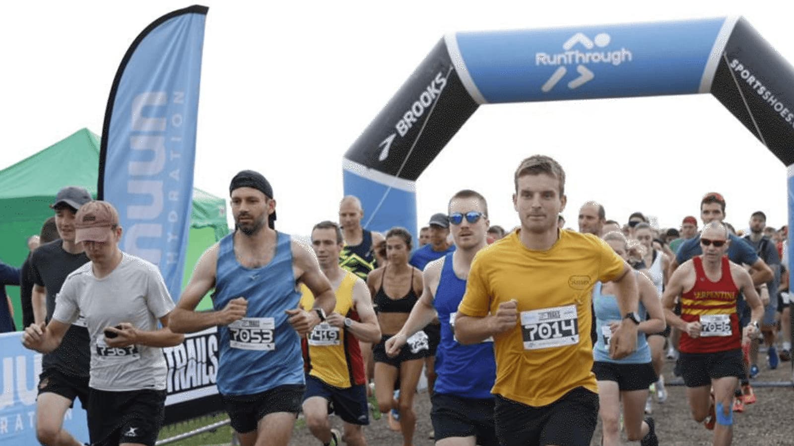 A group of runners in athletic gear start a race under an inflatable RunThrough arch. Some wear sunglasses and numbered bibs. Spectators and tents are visible in the background.