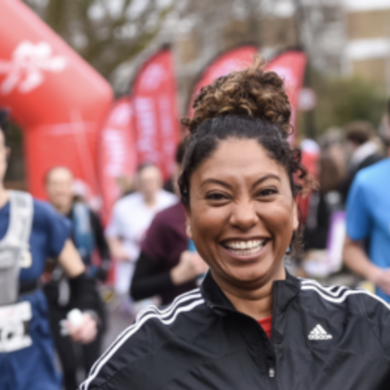 A smiling woman in athletic gear holds a smartphone and a bottle of orange drink while running in a race. Other runners are visible in the background under a red archway with the words "Surrey Half" on it. The atmosphere appears energetic and joyful.